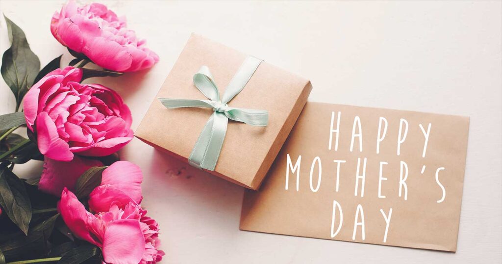 happy mother's day text on craft card and pink peonies bouquet with gift box on rustic white wooden background in light.